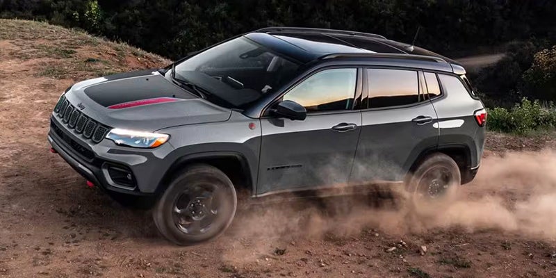 A dark gray Jeep Compass with black roof and black-and-red hood detailing tears up a dirt road.