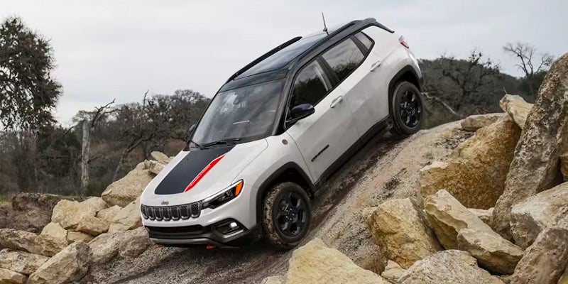 A sporty-looking white Jeep Compass with a black roof and black-and-red hood detailing navigates a steep, rocky hill, heading downhill.