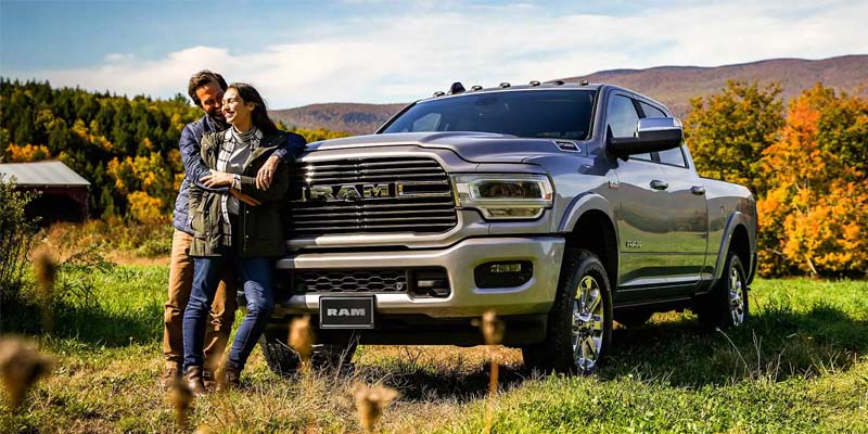 An image of a couple leaning against the front of their grey RAM 2500 in an embrace. The truck is parked on the grass in a field backed by an autumn forest.