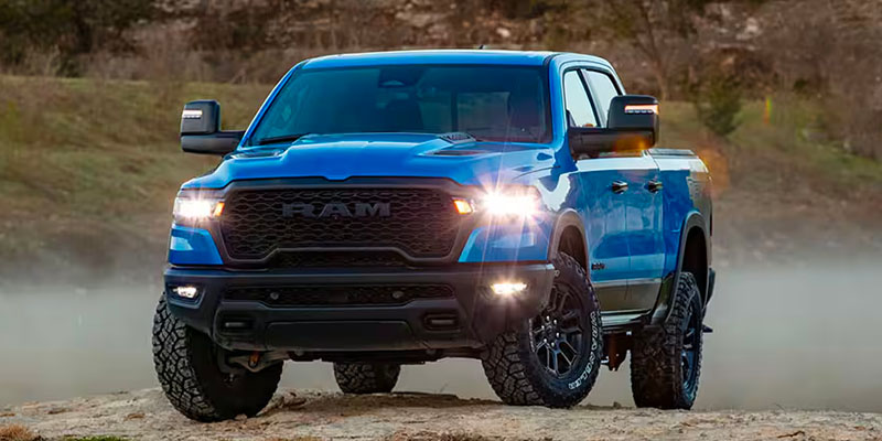 An image of a blue RAM 1500 with its headlights on, sitting on a rocky field backed by a scrubgrass-covered mountain at dusk.